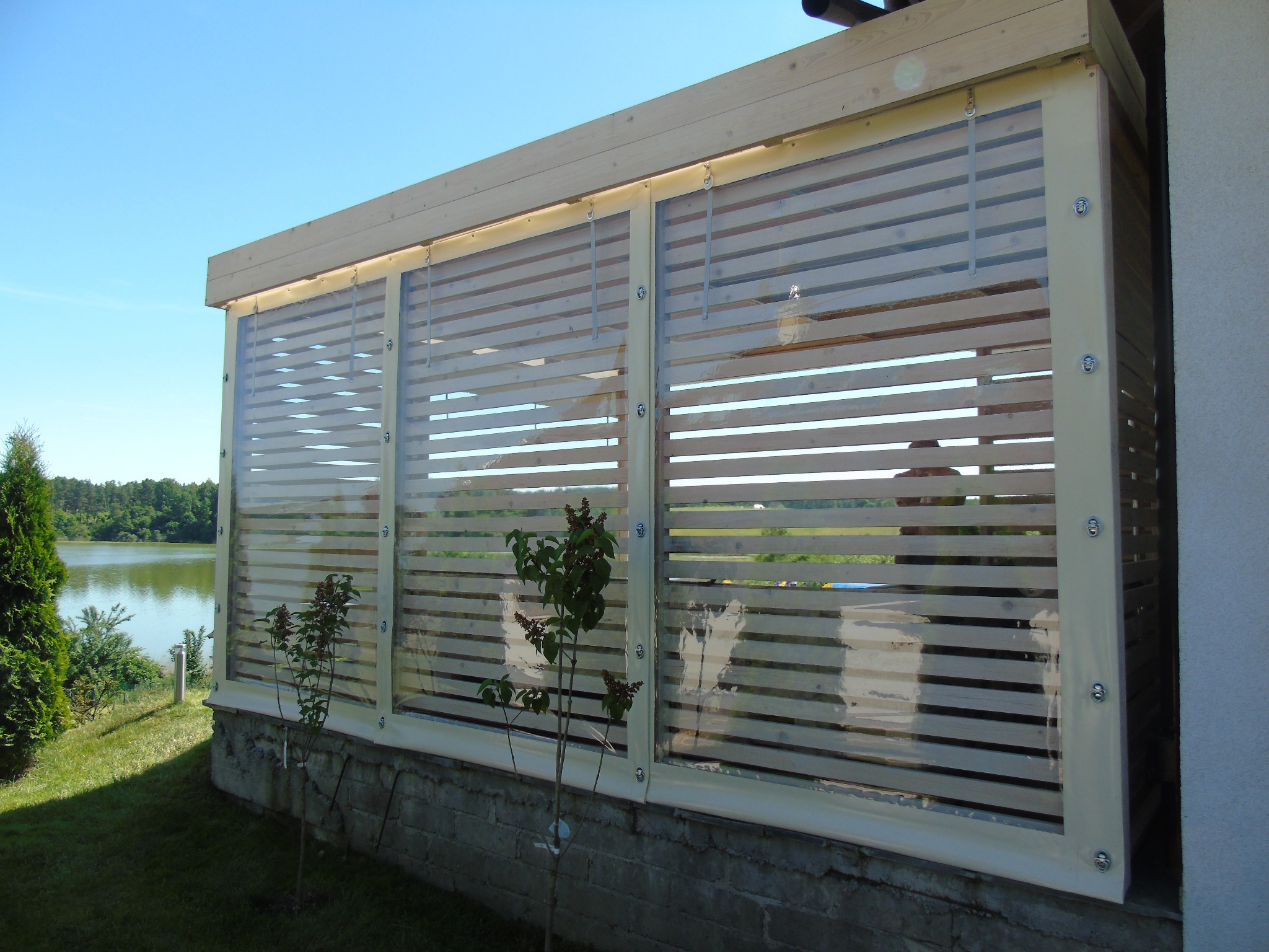 Przezroczyste osłony przeciwwiatrowe z poziomymi listwami na tarasie z widokiem na jezioro, zamocowane śrubami do drewnianej konstrukcji.