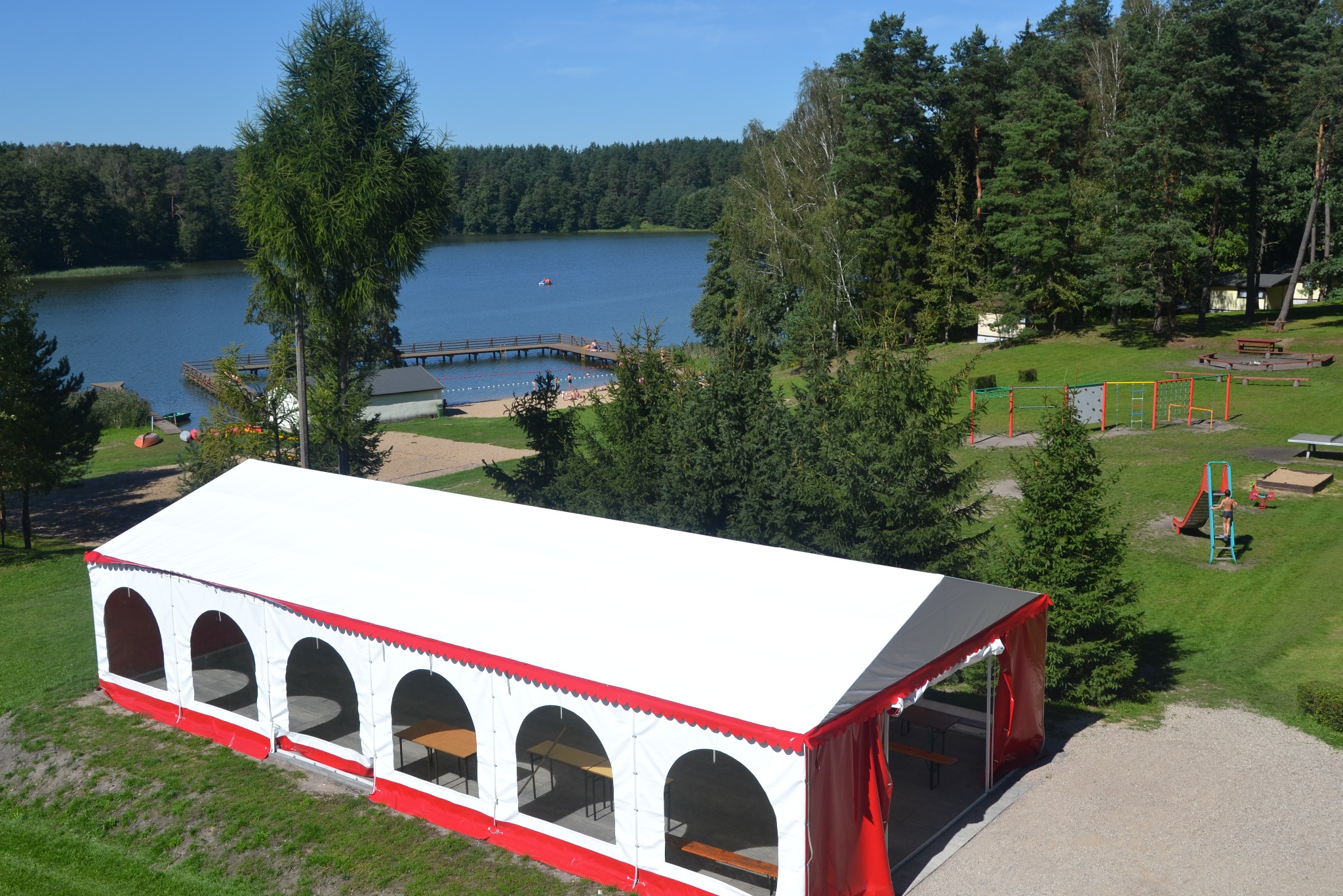 Biały namiot plenerowy z czerwonymi akcentami, widok z góry na tle jeziora, lasu i placu zabaw.
