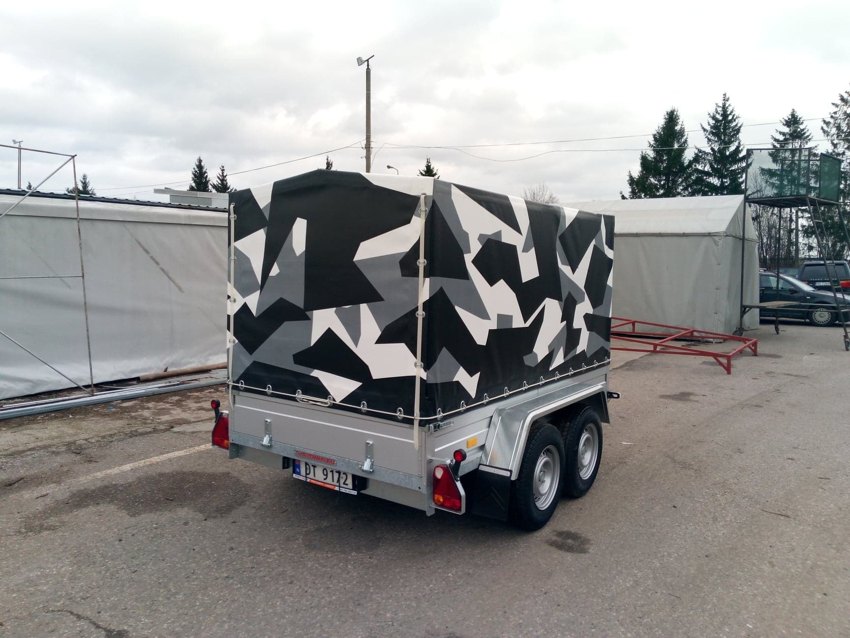 Przyczepa z pokrowcem w kamuflażu szaro-biało-czarnym, widok z tyłu, na tle szarego parkingu i namiotów.