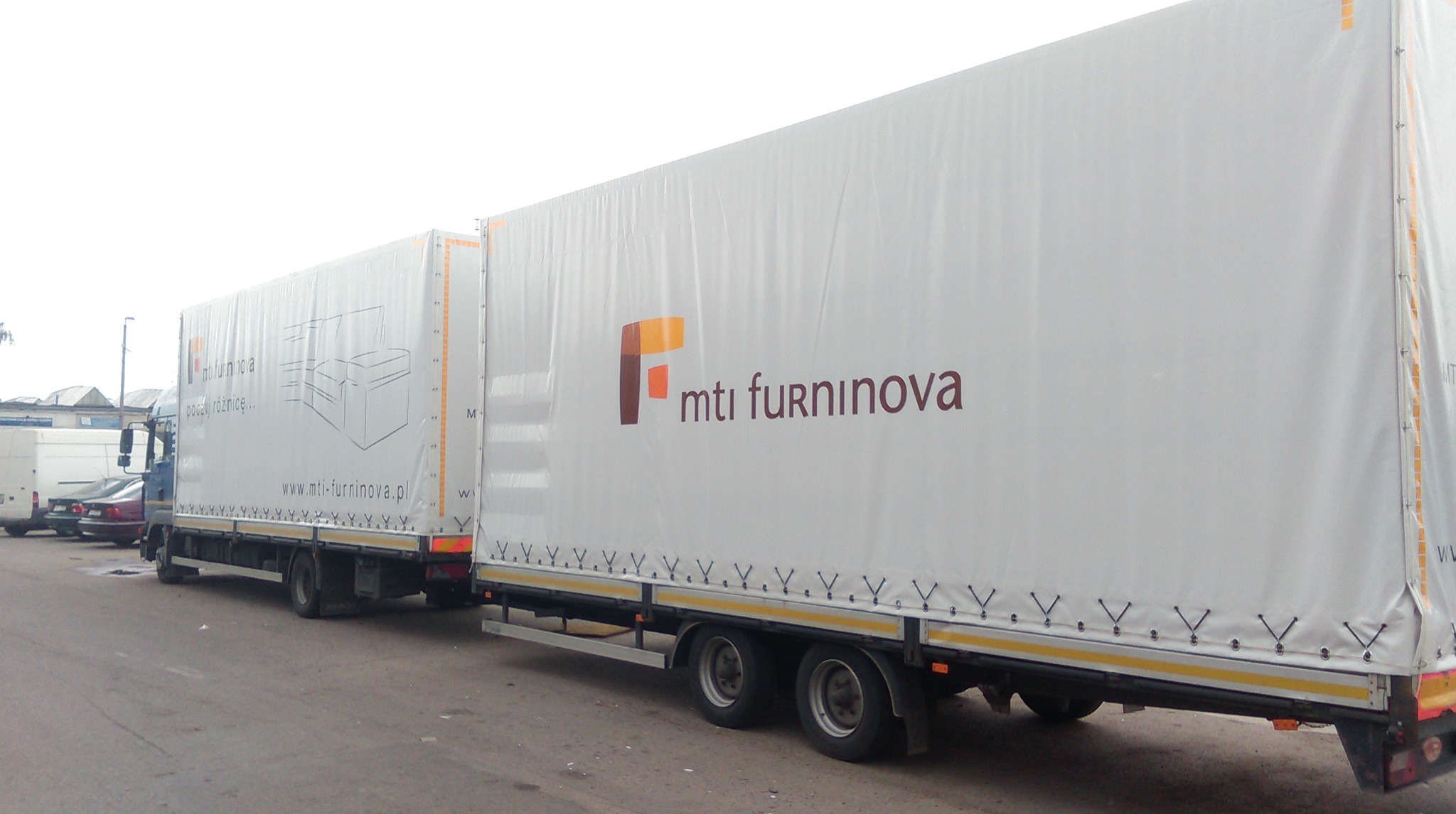 Długi ciągnik siodłowy z naczepą, pokryty białą plandeką z logo MTI Furninova, prezentujący meble tapicerowane w formie grafiki na boku.
