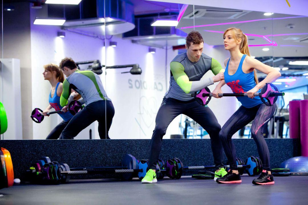 Mężczyzna w szarej koszulce z zielonymi rękawami pomaga kobiecie w niebieskim topie ćwiczyć z obciążeniem w siłowni z lustrami i fioletowym oświetleniem.