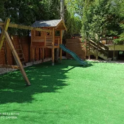 Przydomowy plac zabaw z drewnianym domkiem na palach ze zjeżdżalnią, huśtawką i sztuczną trawą.