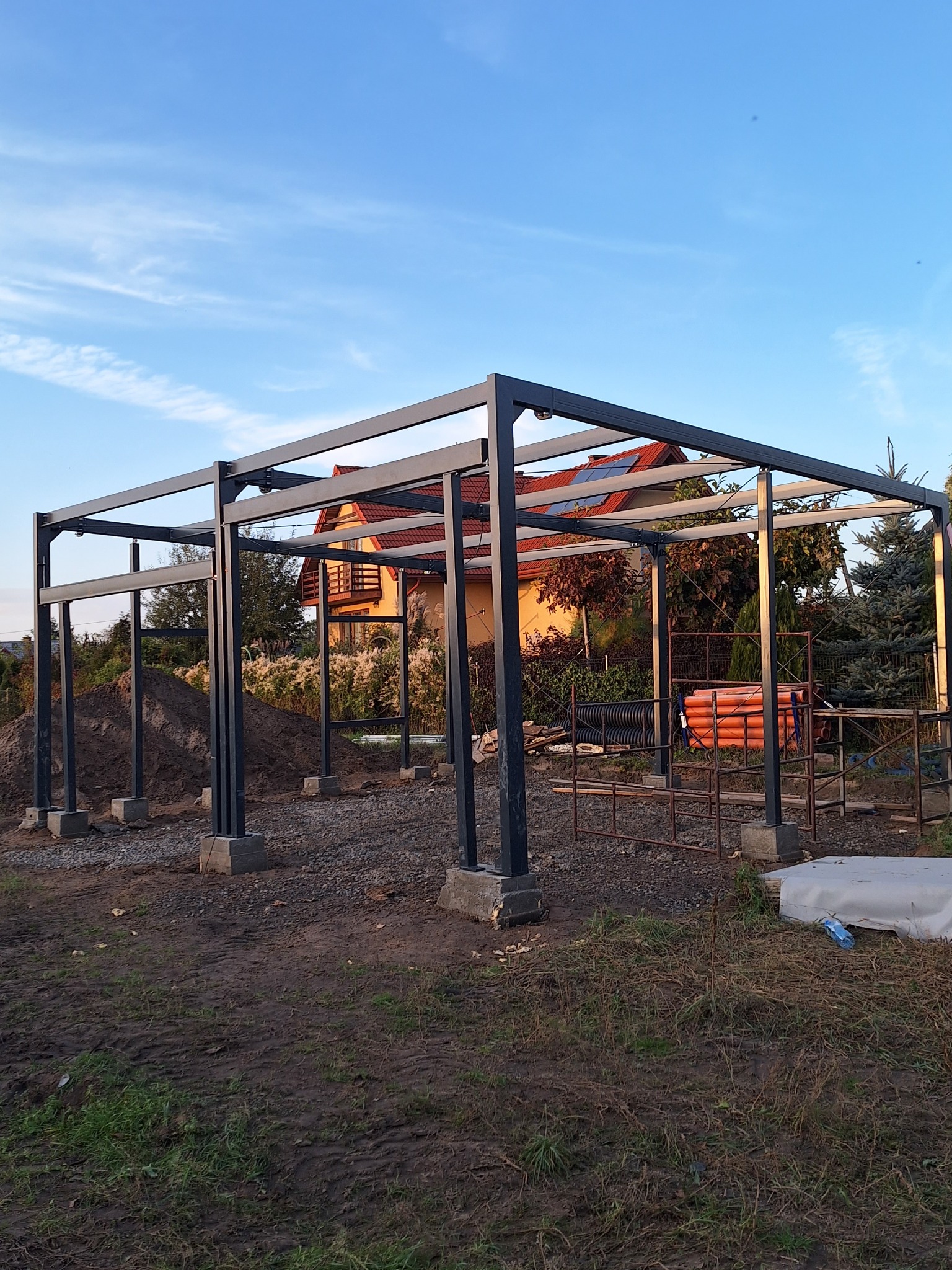 Metalowa konstrukcja z fundamentami na działce, w tle dom z czerwoną dachówką i błękitne niebo. Widoczne rury i materiały budowlane.