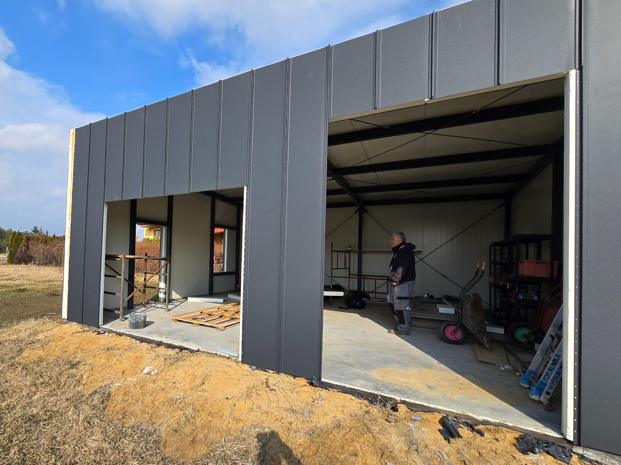 Konstrukcja modułowa w trakcie budowy: szare panele ścian, widoczne elementy konstrukcyjne, człowiek wewnątrz. Częściowo wykończony budynek z otwartymi wrotami.