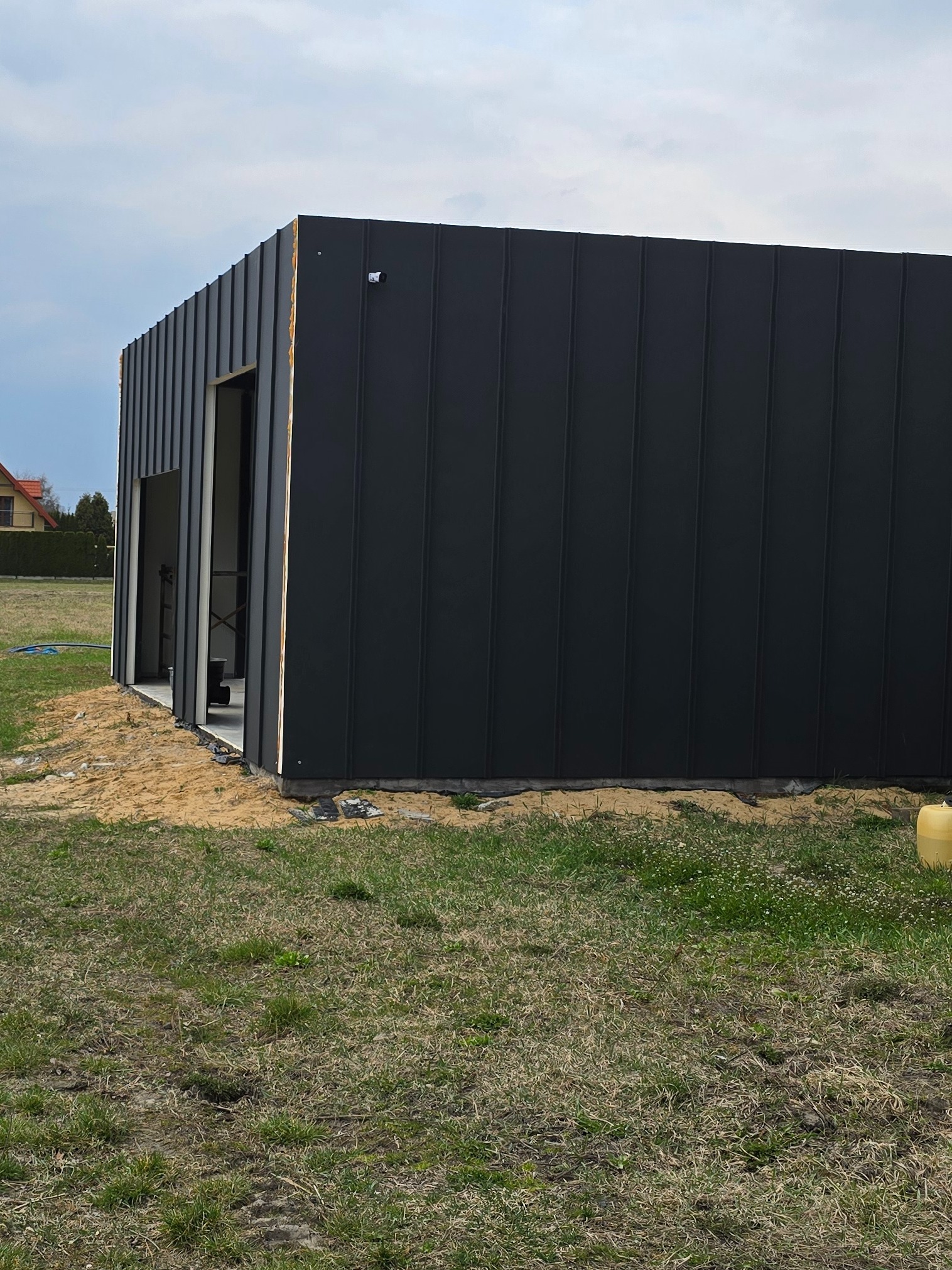Ciemnoszara bryła domu modułowego w trakcie budowy, z widocznymi otworami okiennymi i drzwiowymi, na tle zielonej trawy i błękitnego nieba.