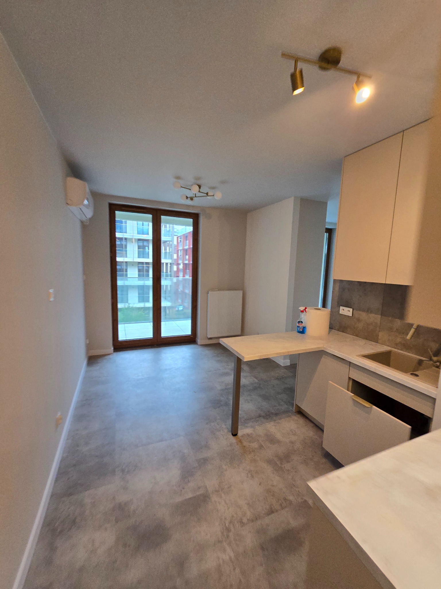 Jasne wnętrze mieszkania po remoncie: szare panele, okno balkonowe, aneks kuchenny z blatem i szafkami. Widok na budynek z zewnątrz.