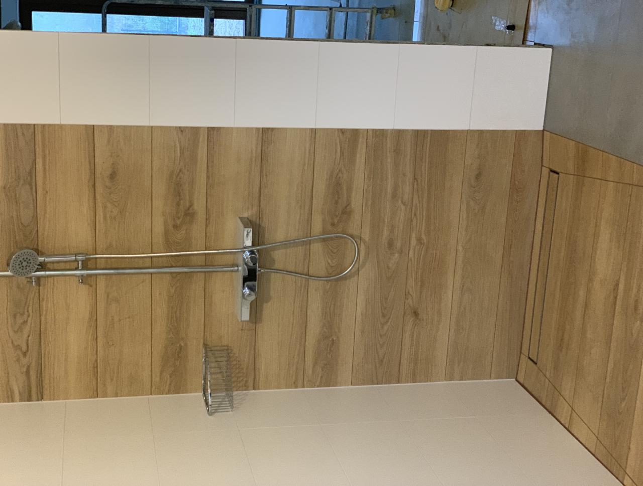 Wykończona łazienka w Warszawie z drewnopodobnymi panelami ściennymi, białymi płytkami u góry i zamontowanym zestawem prysznicowym ze słuchawką. Widoczny fragment drabiny w górnej części zdjęcia...