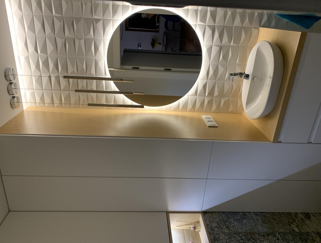 Nowoczesna łazienka z umywalką nablatową, okrągłym lustrem z podświetleniem LED i dekoracyjną ścianą z białych, trójwymiarowych płytek. Widoczne minimalistyczne oświetlenie sufitowe na cienkich...