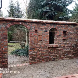 Ceglana ściana z dwoma łukowymi otworami i jednym prostokątnym, w ogrodzie, z widocznymi śladami zaprawy i różnicami w kolorze cegieł.