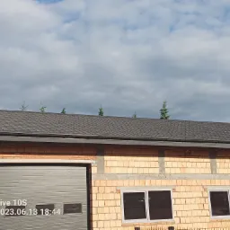 Budynek w trakcie budowy z widocznym nowym dachem z ciemnej dachówki, jasną cegłą i szarą bramą garażową, z młodymi drzewkami iglastymi wyrastającymi ponad linię dachu.