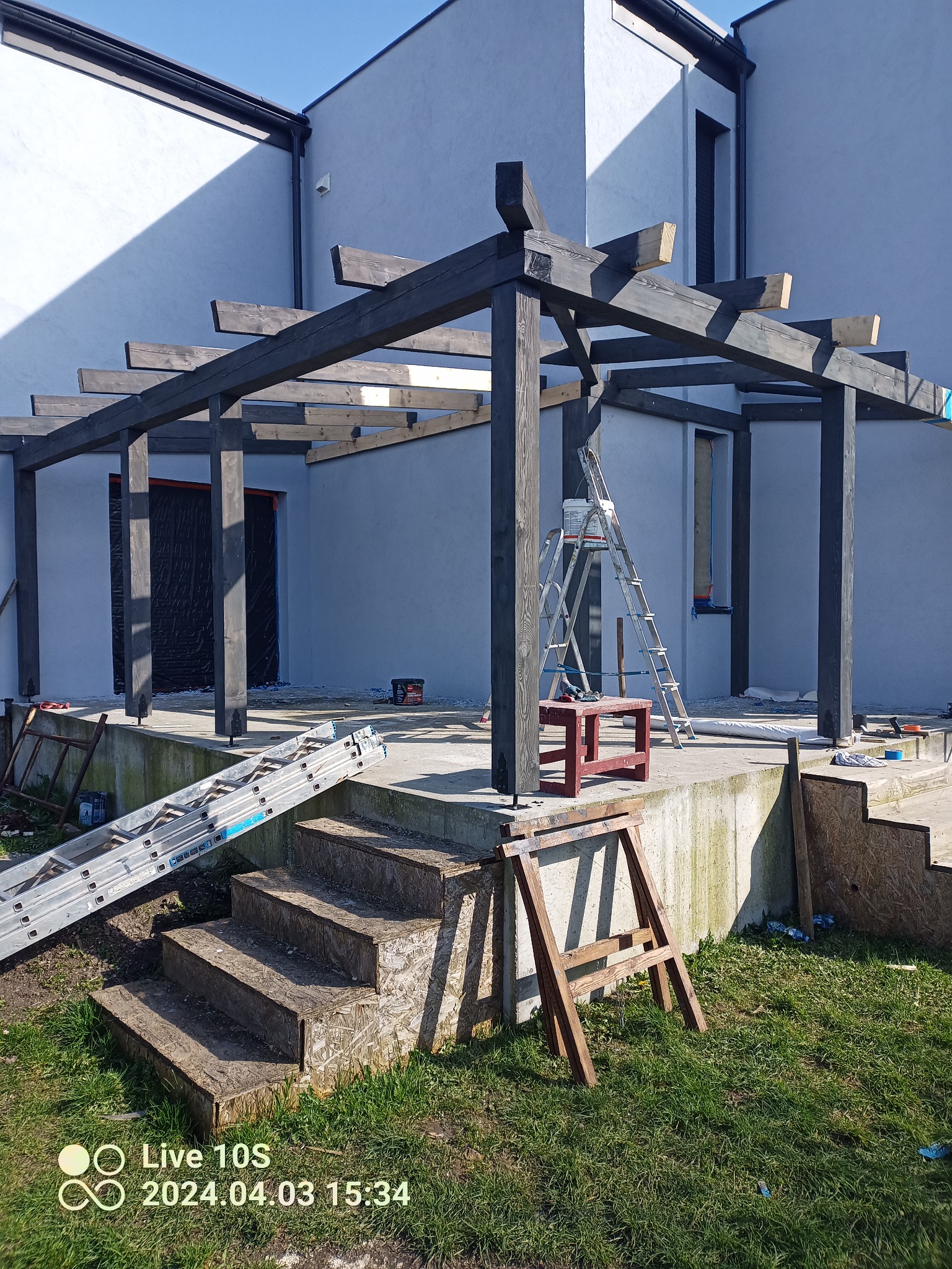 Montaż drewnianej pergoli przy nowoczesnym domu, widoczne rusztowania, drabiny i narzędzia, etap budowy.