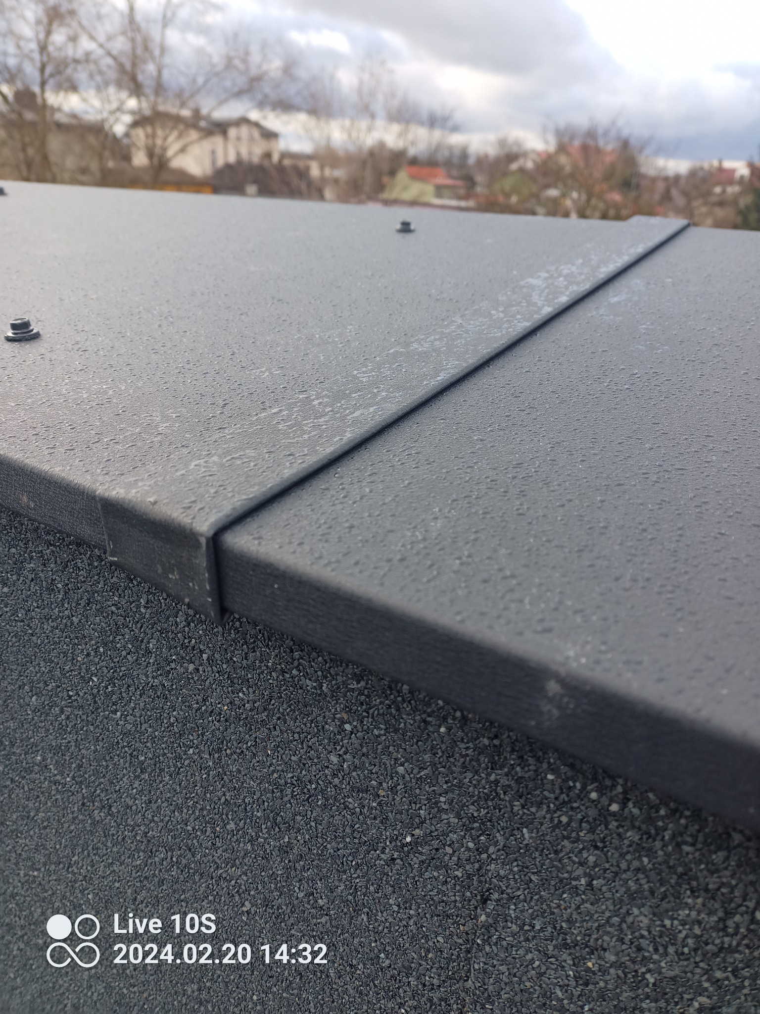 Fragment czarnego, metalowego dachu z widocznymi kroplami wody, połączonego z pokryciem bitumicznym, z widokiem na zabudowania w oddali i pochmurne niebo.