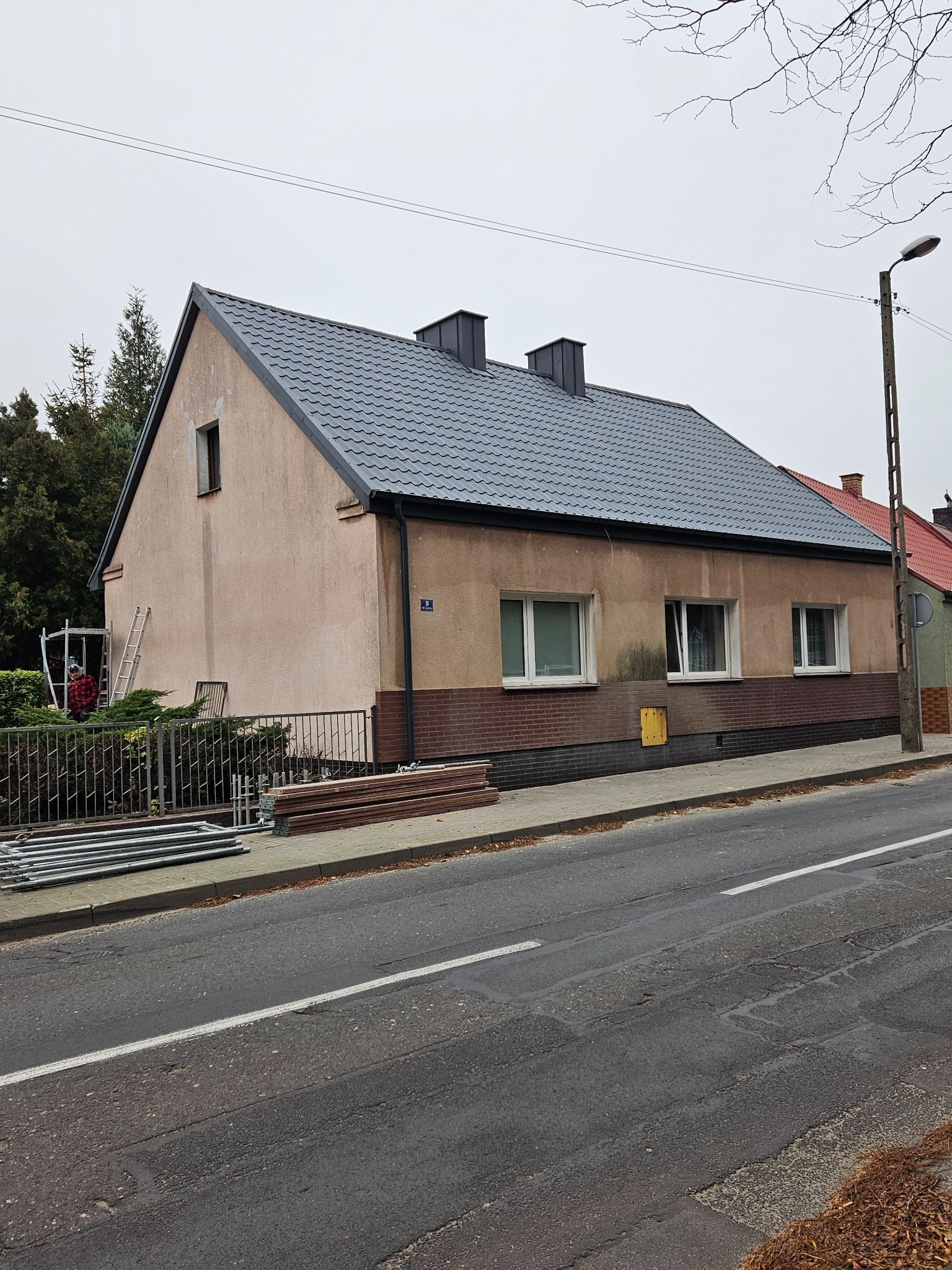 Dom jednorodzinny z nowym, ciemnoszarym dachem z blachodachówki, widoczne kominy, drabina oparta o ścianę, rusztowania rozłożone na chodniku przed domem, ulica na pierwszym planie.