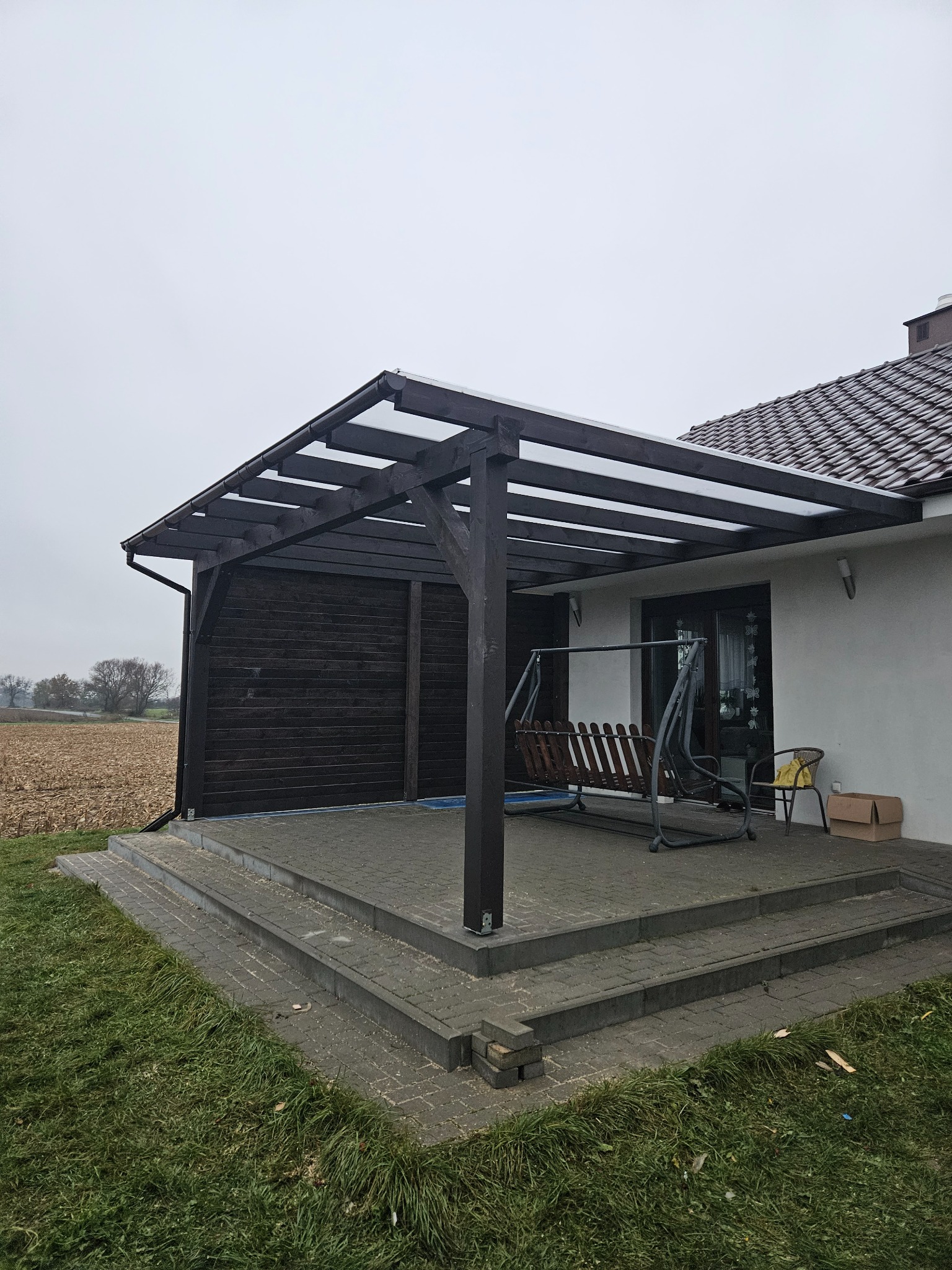 Zadaszony taras z drewnianą konstrukcją, huśtawką i widokiem na pole w pochmurny dzień.