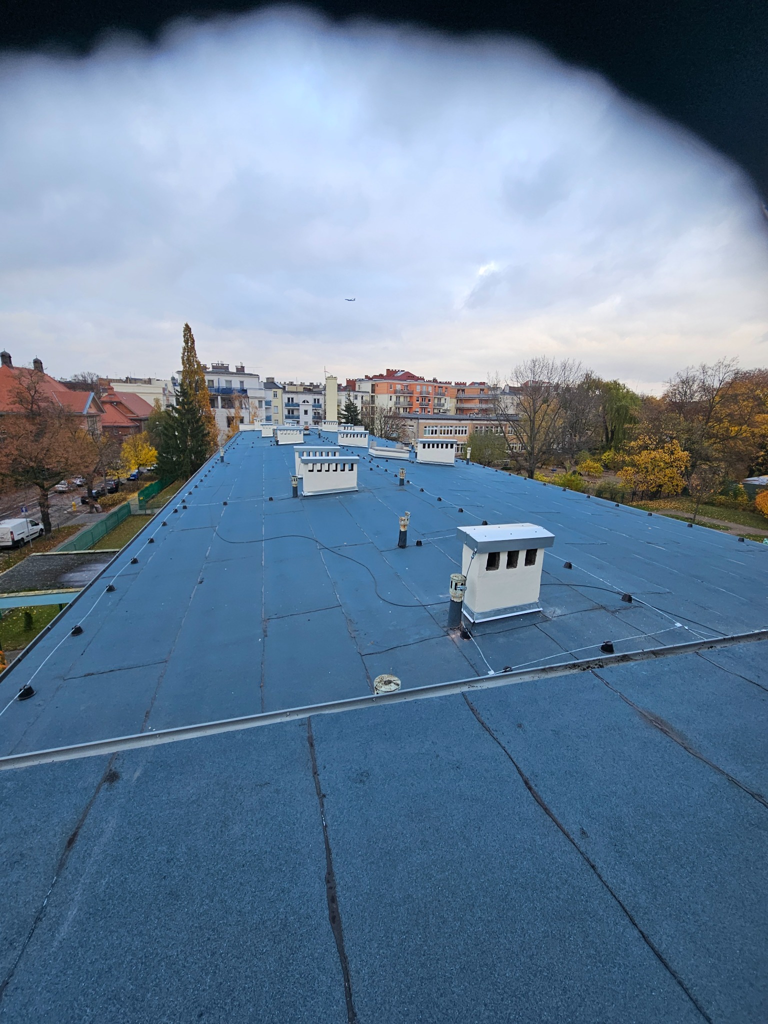 Niebieski dach budynku z kominami na tle miejskiego krajobrazu i pochmurnego nieba. Widoczny samolot w oddali. Dach pokryty papą.
