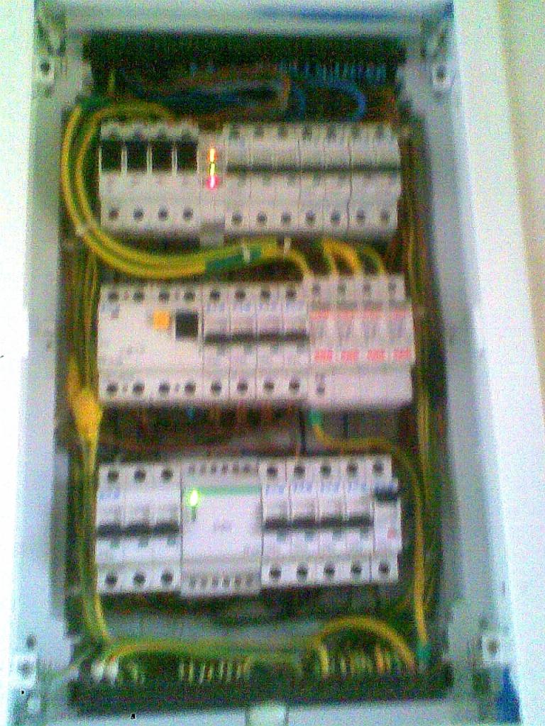 Otwarta skrzynka elektryczna z trzema rzędami bezpieczników, widoczne przewody w kolorach żółto-zielonym, jeden bezpiecznik oznaczony czerwoną diodą, drugi zieloną.