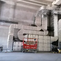 Montaż systemu wentylacji przemysłowej z użyciem podnośnika Heron, widoczne izolowane kanały wentylacyjne i pracownik w kasku.