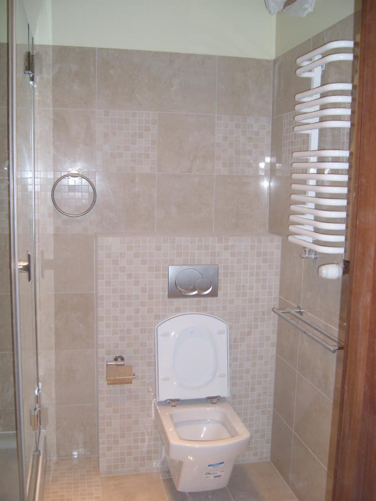 Wykończona łazienka z beżowymi płytkami, wiszącą toaletą z otwartą klapą, grzejnikiem drabinkowym i szklaną kabiną prysznicową.