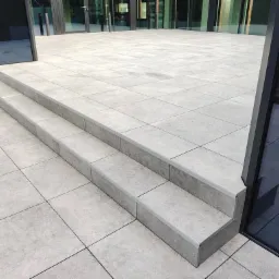 Jasnoszara, gładka nawierzchnia tarasu z dwustopniowymi schodami prowadzącymi do przeszklonego wejścia budynku.