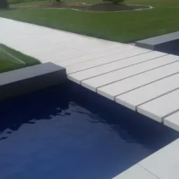 Nowoczesny taras z białych płyt przechodzący nad basenem, z systemem nawadniania trawnika w tle.