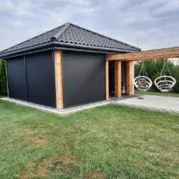Nowoczesna altana z czarnymi, zwijanymi osłonami przeciwsłonecznymi, drewnianą konstrukcją i wiszącymi fotelami na tle zielonego trawnika i pochmurnego nieba.