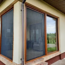 Zewnętrzna, plisowana ochrona okienna w kolorze grafitowym zamontowana na dużych, brązowych oknach tarasowych, widok z zewnątrz na tle zadbanego ogrodu.