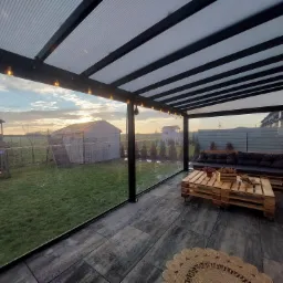 Zadaszony taras z przezroczystymi roletami screen zip, meble z palet, widok na ogród i zachodzące słońce.