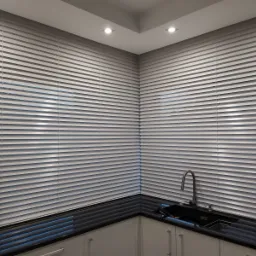 Jasne, poziome listwy osłonowe okienne zamontowane w rogu kuchni z czarnym blatem i białymi szafkami, widoczne fragmenty okna i zwisające sznurki regulacyjne.