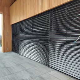 Zewnętrzne żaluzje poziome, lamele w kolorze grafitowym, zamontowane na tarasie z drewnianą konstrukcją, widoczny fragment sterowania elektrycznego.