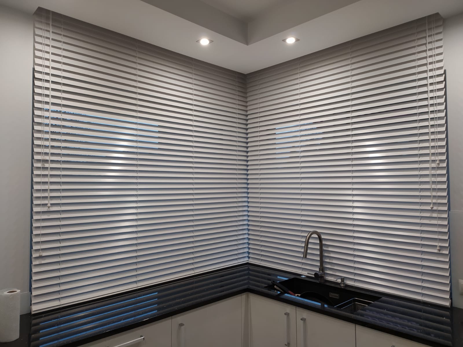 Jasne, poziome listwy osłonowe okienne zamontowane w rogu kuchni z czarnym blatem i białymi szafkami, widoczne fragmenty okna i zwisające sznurki regulacyjne.