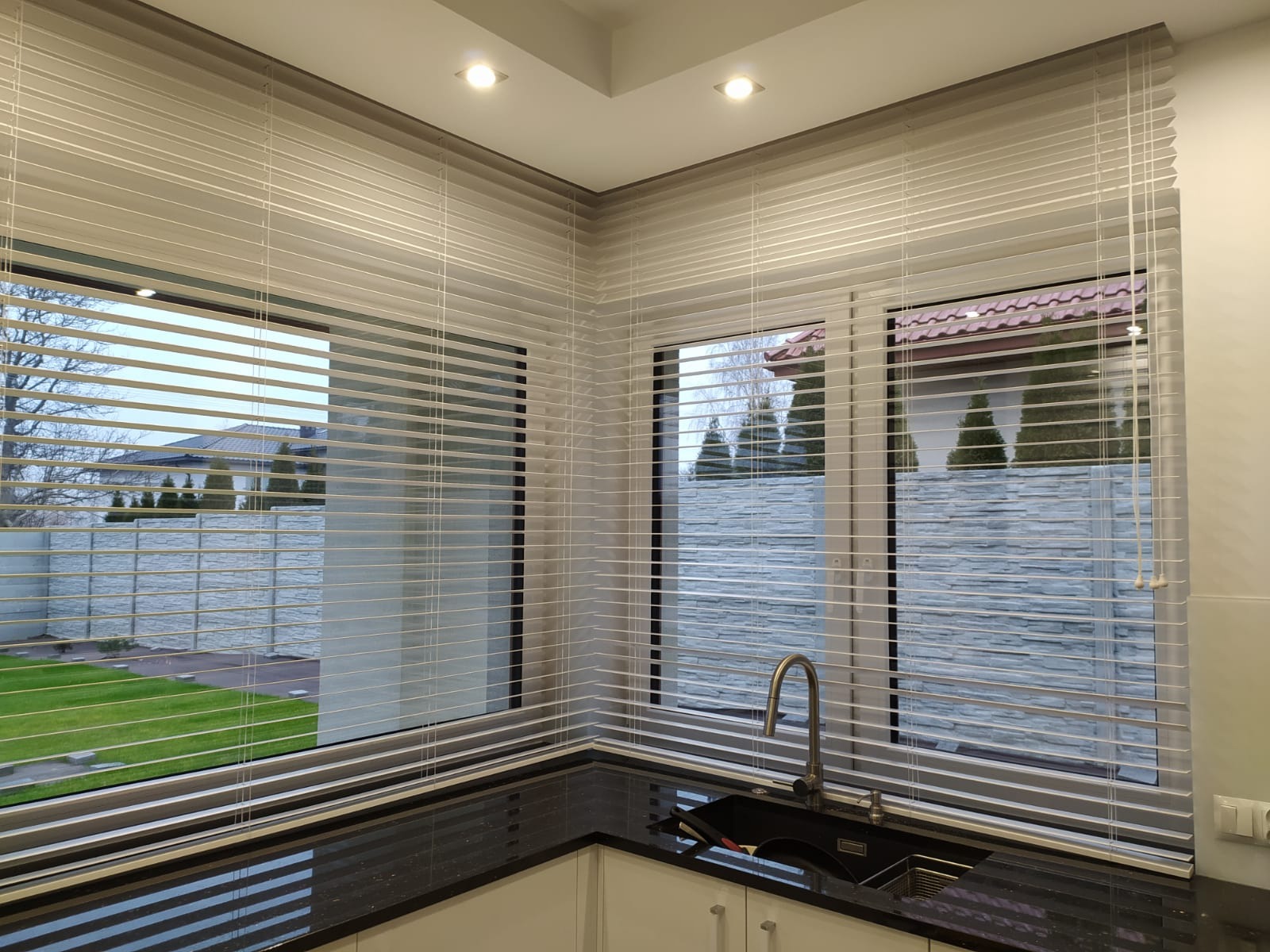 Jasne żaluzje drewniane 50mm zamontowane w narożnym oknie kuchni z widokiem na ogród i dom.