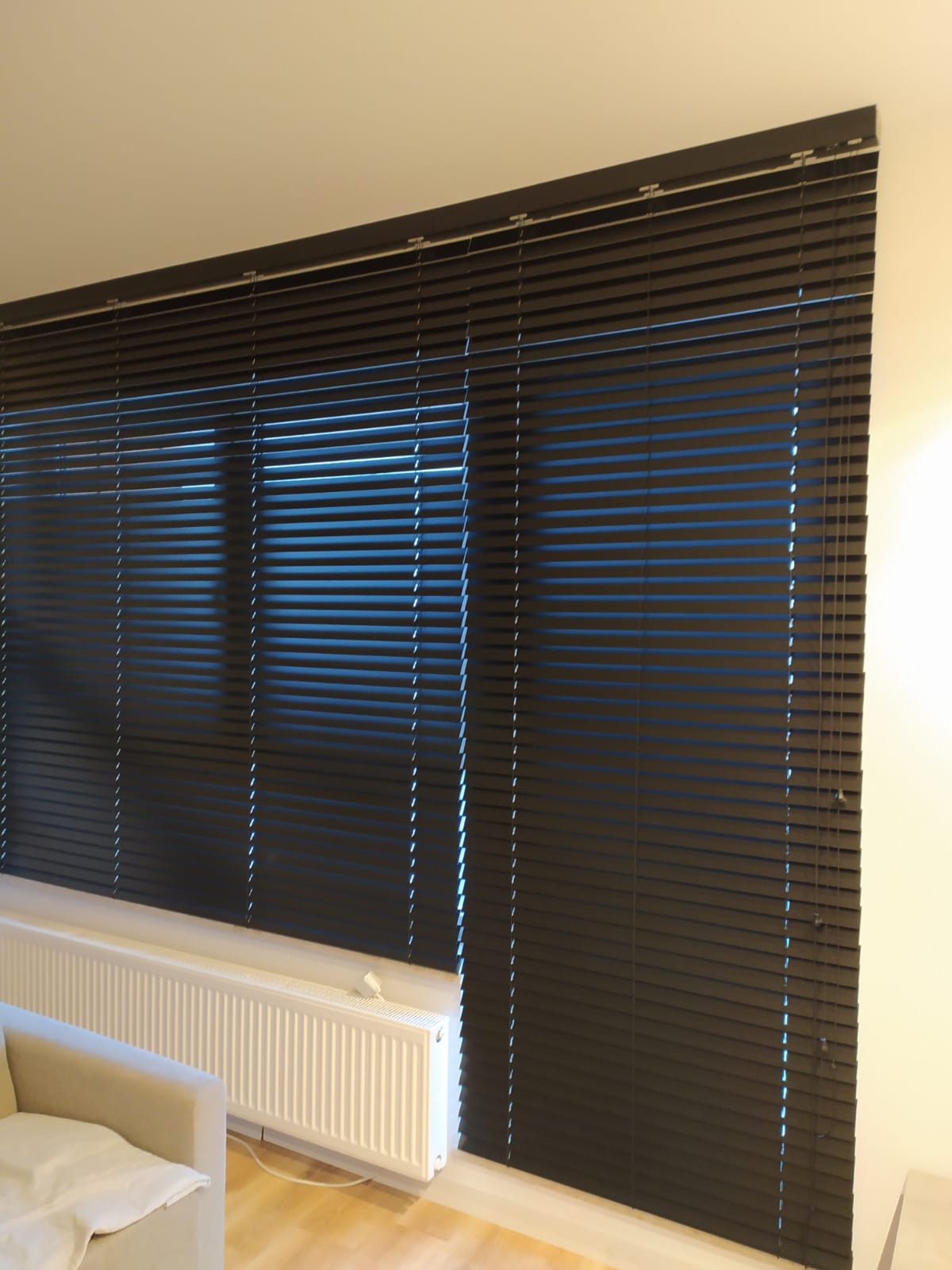 Ciemne, poziome listwy osłaniające okno, prawdopodobnie żaluzje, z widocznym fragmentem grzejnika i jasnej sofy w tle.