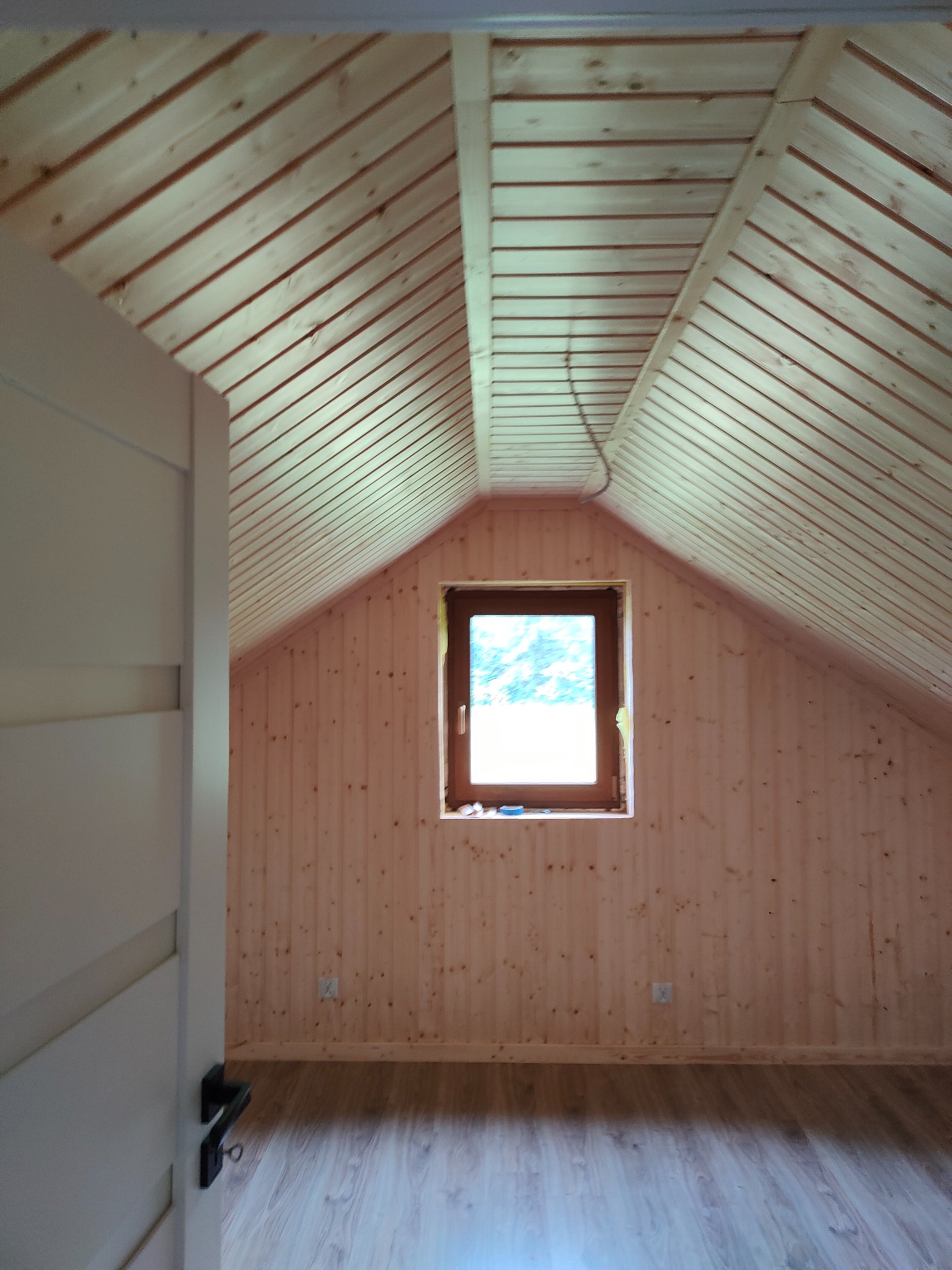 Poddasze wykończone boazerią z jasnego drewna, z oknem w ramie z ciemniejszego drewna i widoczną instalacją elektryczną pod sufitem, drzwi wejściowe z czarną klamką z lewej strony.