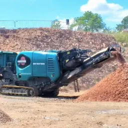 Niebieska mobilna kruszarka METROTRAK przerabia gruz na placu budowy, tworząc pryzmę drobnego kruszywa; w tle hałda gruzu i błękitne niebo.
