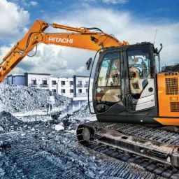 Pomarańczowa koparka Hitachi nabiera ziemię z wykopu na tle budowanego budynku i błękitnego nieba z chmurami.