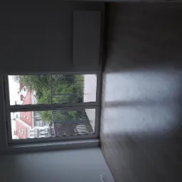 Pusty pokój z panelami podłogowymi w odcieniach szarości, widok na balkon i budynki zza okna.