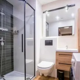 Nowoczesna łazienka z kabiną prysznicową typu walk-in z czarnymi profilami, podwieszaną toaletą, drewnianą szafką pod umywalką i dużym lustrem z oświetleniem punktowym.