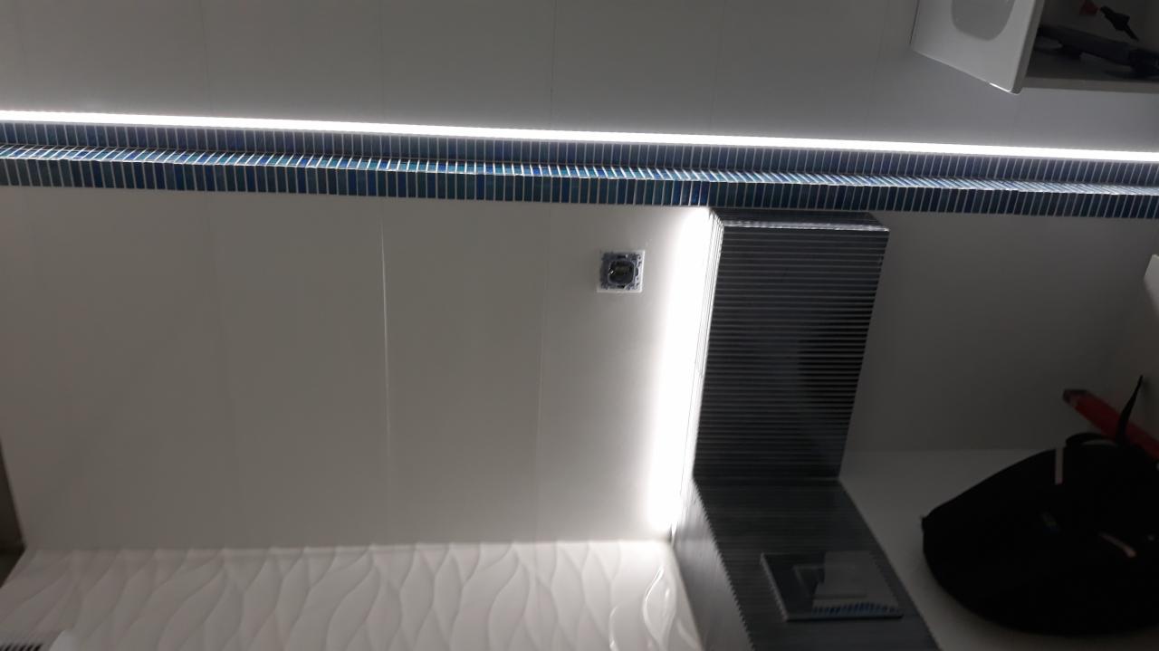 Nowoczesna łazienka z białymi płytkami falistymi na dole ściany, niebieską mozaiką pod sufitem i oświetleniem LED, widoczne gniazdko elektryczne oraz fragment czarnej torby.