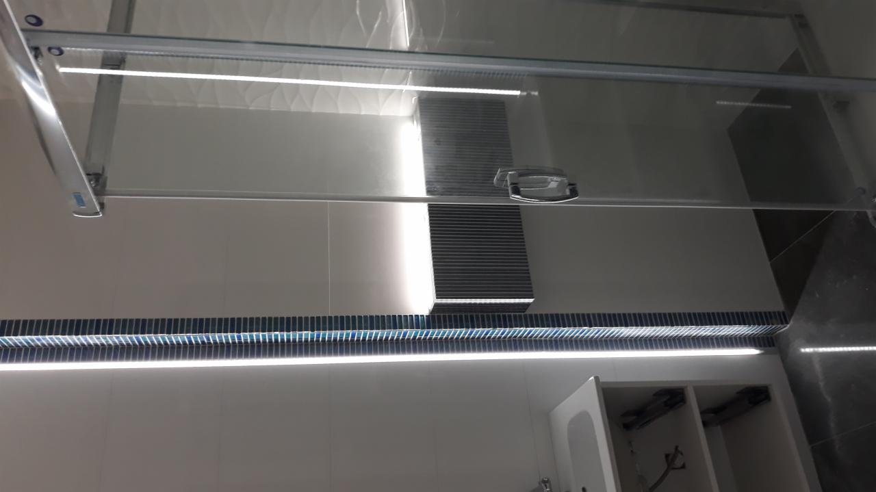 Szklana kabina prysznicowa z przesuwnymi drzwiami, oświetleniem LED i mozaiką w odcieniach niebieskiego, zamontowana w nowoczesnej łazience z białymi płytkami.