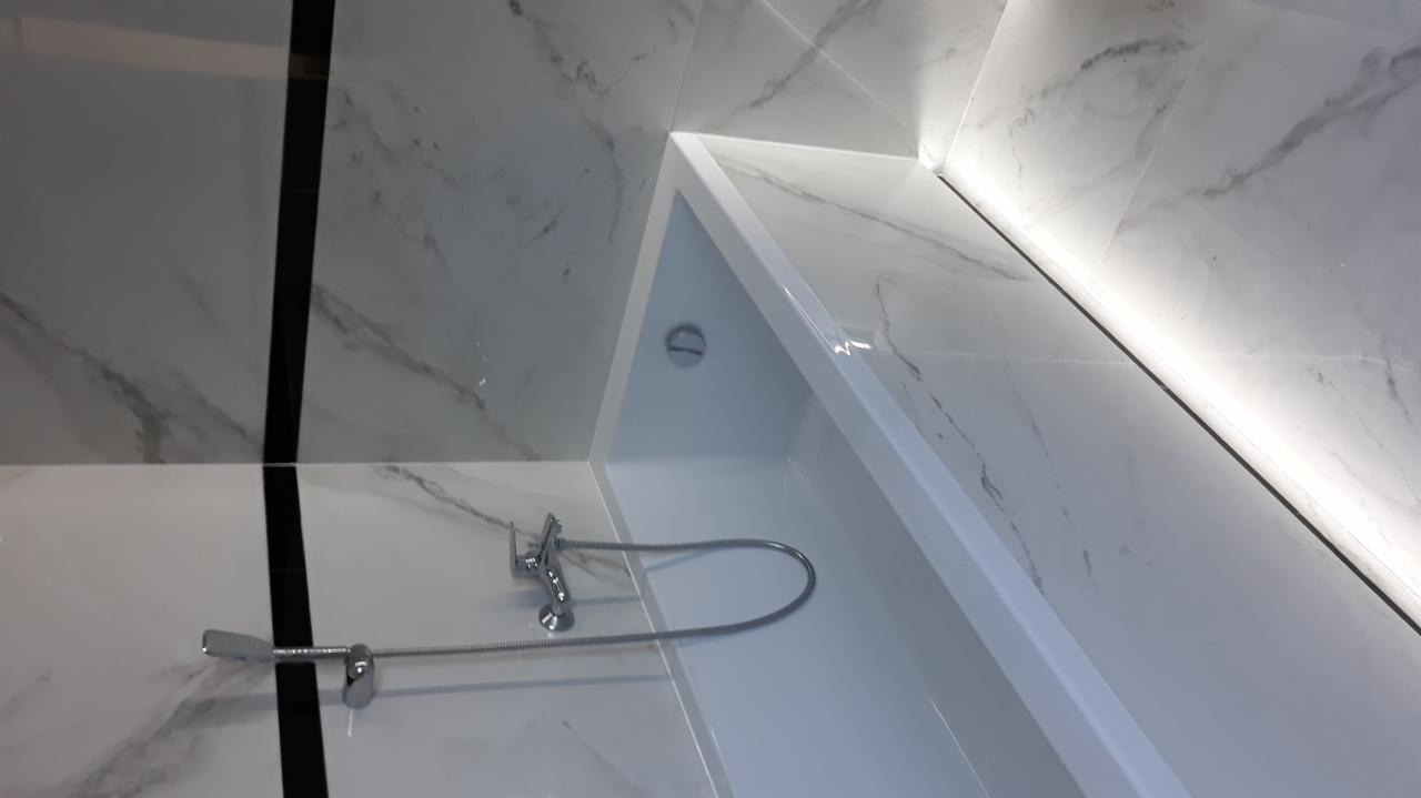 Nowoczesna łazienka z białą wanną, armaturą i marmurowymi płytkami z czarnym akcentem, oświetlenie LED wzdłuż krawędzi.