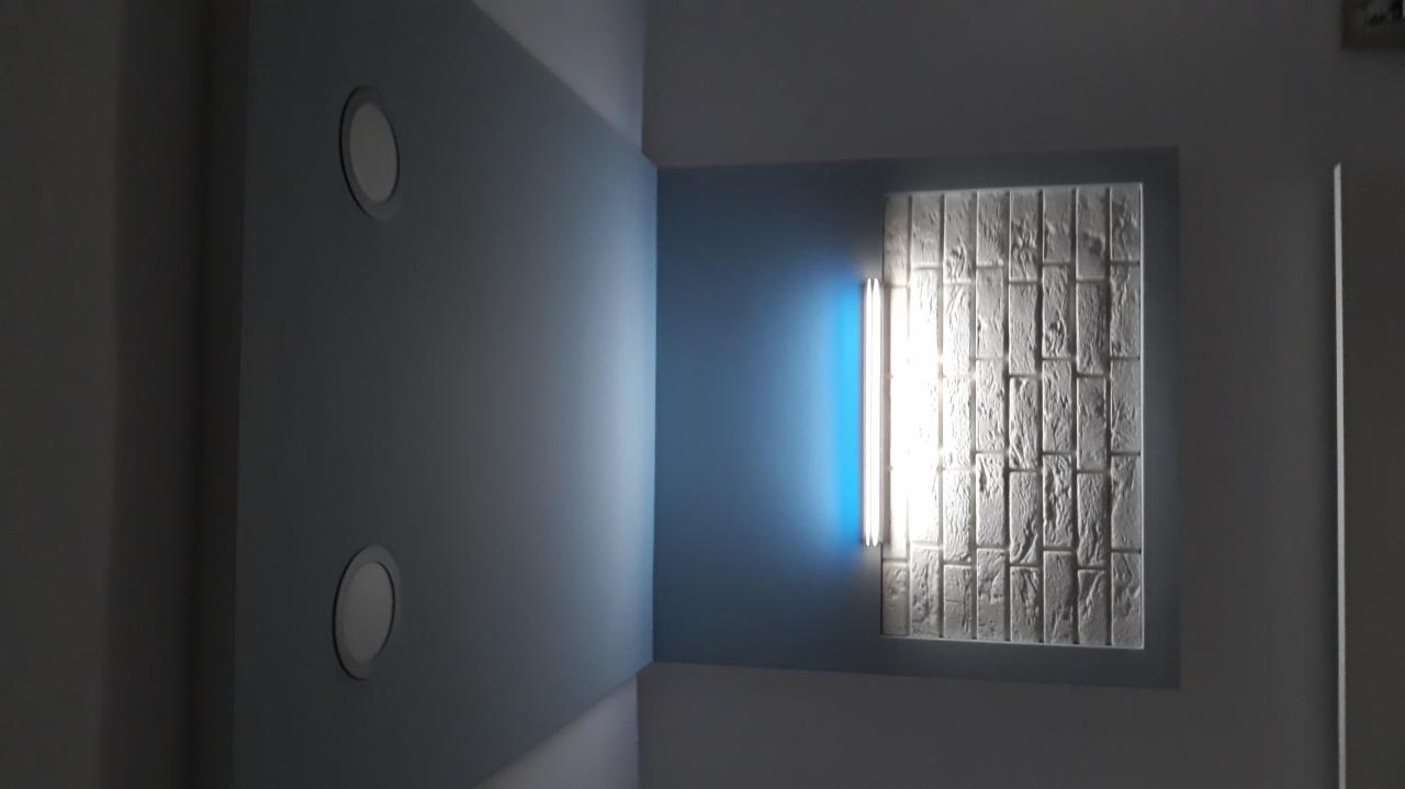 Narożnik pokoju z niebieskimi ścianami, dwoma okrągłymi lampami w suficie i dekoracyjną ścianą z białej cegły podświetloną pionową lampą.