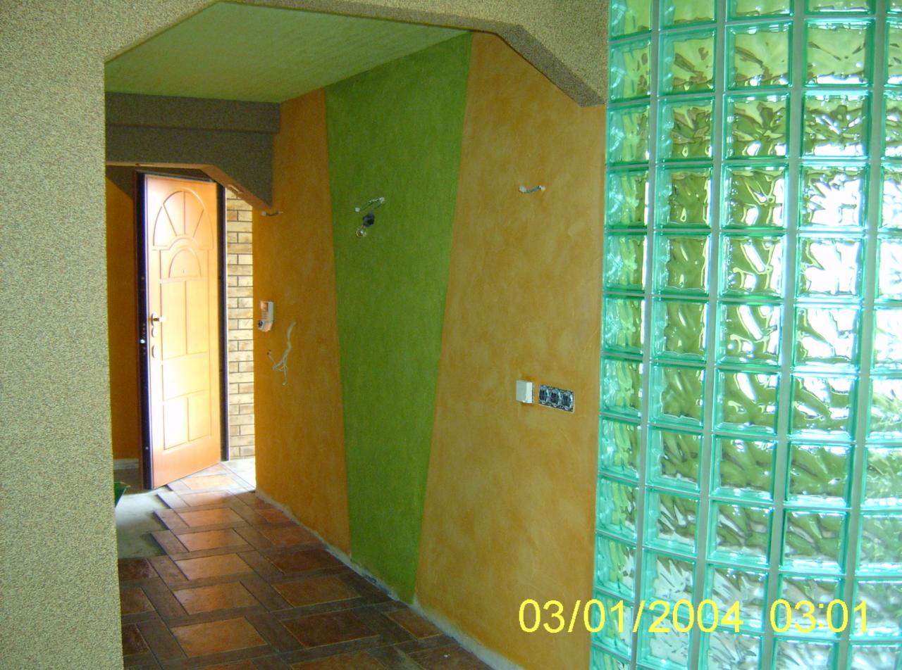 Korytarz z dekoracyjną ścianą w odcieniach zieleni i pomarańczy, wykończony panelami z luksferów, widoczne przygotowania do montażu gniazdek elektrycznych i otwarte drzwi wejściowe z ceglaną ścianą...