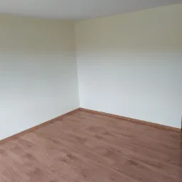 Pusty pokój po remoncie z panelami podłogowymi w odcieniach brązu i beżowymi ścianami, widoczne wystające kable z sufitu.