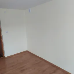 Pomalowane na jasnożółto ściany i brązowa podłoga z paneli w rogu pokoju, widoczny fragment drzwi i kratka wentylacyjna.