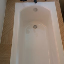 Piotr Pilecki - Biała wanna z czarną armaturą, obudowana płytkami w odcieniach beżu, ułożonymi we wzór jodełki. Widok z góry, minimalistyczny design.