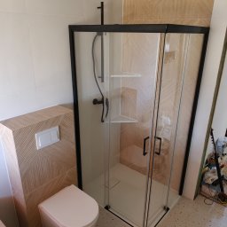 Piotr Pilecki - Nowoczesna łazienka po remoncie: kabina prysznicowa z czarną ramą, podwieszana toaleta z drewnopodobną obudową i wzorzyste płytki podłogowe.