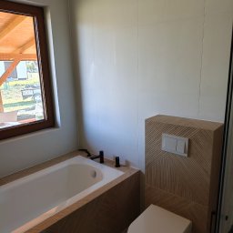 Piotr Pilecki - Nowoczesna łazienka z wanną obudowaną drewnopodobną płytką i podwieszaną toaletą. Białe płytki na ścianach, okno z widokiem na zewnątrz. Czarna armatura.