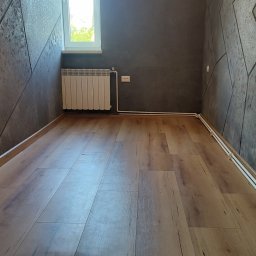 Piotr Pilecki - Pokój po remoncie: panele podłogowe w odcieniach drewna, ściany z dekoracyjną szarą strukturą, biały grzejnik pod oknem. Widok na wnętrze z perspektywy podłogi.