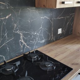 Piotr Pilecki - Czarny marmurkowy backsplash z płytkami nad blatem i płytą gazową Amica. Ujęcie z góry, widoczne szafki w odcieniach drewna i szarości.