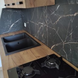 Piotr Pilecki - Nowoczesna kuchnia z czarnym, marmurkowym panelem nad blatem, drewnianymi szafkami i czarnym zlewem. Widoczna płyta gazowa Amica. Ujęcie z góry, prezentujące detale wykończenia.